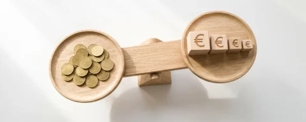 Vue en plongée d'une balance en bois clair sur fond blanc, avec des pièces dorées sur le plateau gauche et des cubes en bois gravés de symboles euro de taille décroissante sur le plateau droit, symbolisant l'équilibre entre revenus publicitaires et réduction des coûts ACOS