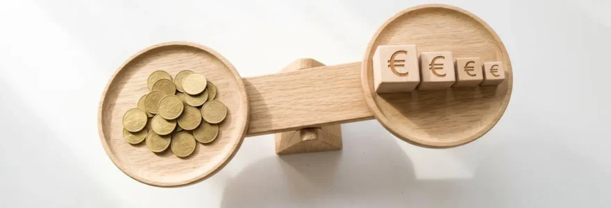Vue en plongée d'une balance en bois clair sur fond blanc, avec des pièces dorées sur le plateau gauche et des cubes en bois gravés de symboles euro de taille décroissante sur le plateau droit, symbolisant l'équilibre entre revenus publicitaires et réduction des coûts ACOS